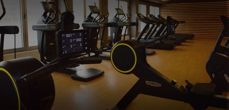 Verschiedene Cardio-Fitnessgeräte stehen aufgereiht vor einer Fensterfront.